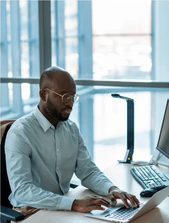 Man in glasses working on a laptop at a desk in a bright office, focused and engaged. A large window provides natural light, creating a calm atmosphere.