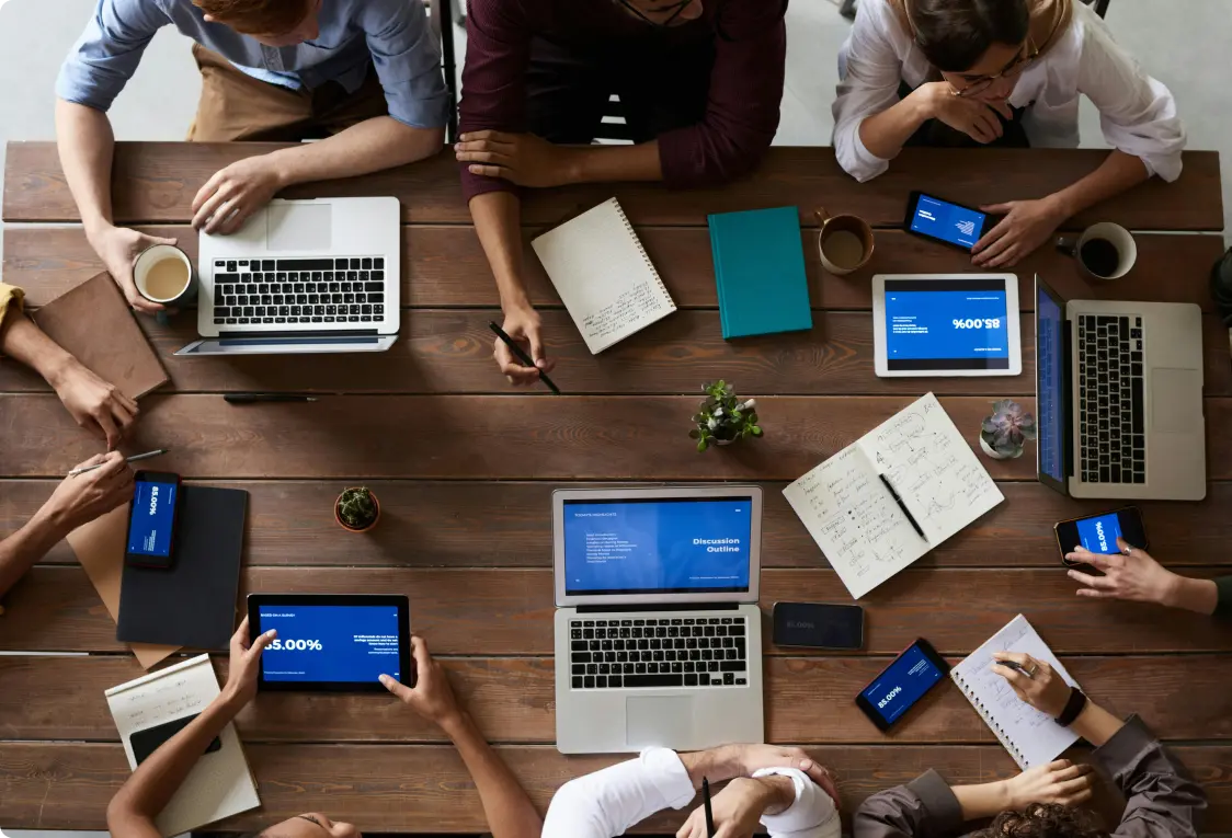 A diverse group of people in a meeting around a wooden table with laptops, phones, and notebooks. The atmosphere is busy and collaborative.